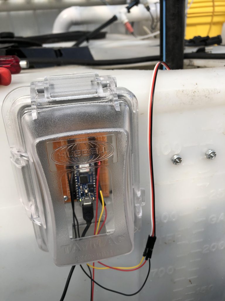A small microcontroller inside a waterproof transparent plastic HUBBELL cover with a thick black cable and thin black, yellow and red cables attached. The microcontroller is secured to the side of a white plastic bin.