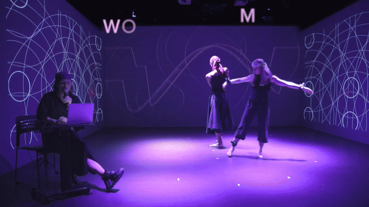 2 dancers move slowly around a small purple-lit stage with various lines and circles projected on 3 surrounding walls. The subtitles WOMAN and MUSIC slowly appear letter-by-letter.